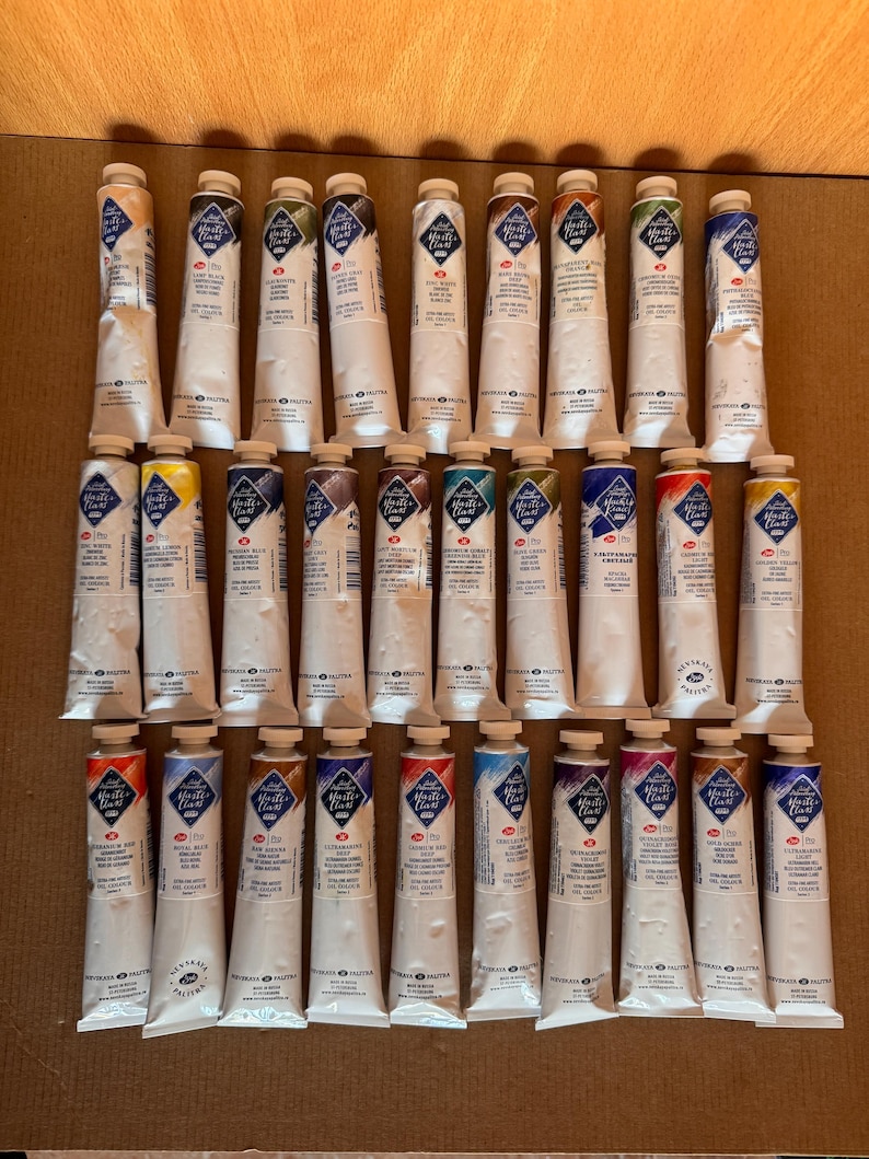 May include: A collection of artist paint tubes, each with a white body and a colored cap. The tubes are arranged in rows, showcasing a variety of colors. The labels feature text and a diamond-shaped logo. The paint tubes are on a brown surface.