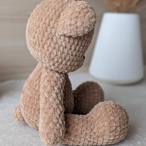 K&ouml;nnte beinhalten: Ein geh&auml;kelter Teddyb&auml;r aus braunem Garn. Der B&auml;r sitzt mit gekreuzten Beinen und geneigtem Kopf.