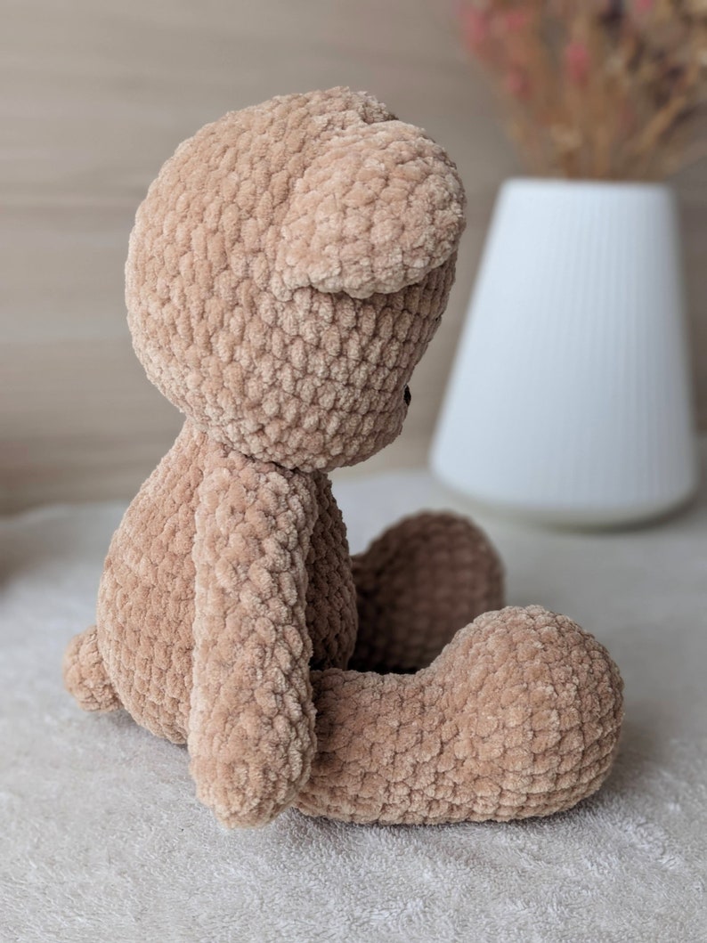 K&ouml;nnte beinhalten: Ein geh&auml;kelter Teddyb&auml;r aus braunem Garn. Der B&auml;r sitzt mit gekreuzten Beinen und geneigtem Kopf.