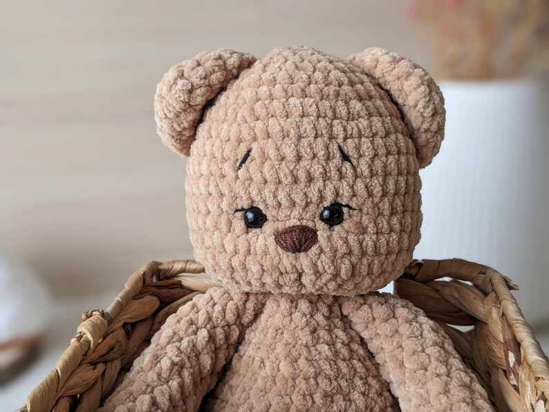 K&ouml;nnte beinhalten: Ein geh&auml;kelter brauner Teddyb&auml;r mit schwarzen gestickten Augen und einer braunen Nase. Der B&auml;r sitzt in einem Weidenkorb.