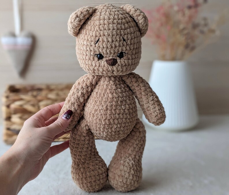 K&ouml;nnte beinhalten: Ein brauner geh&auml;kelter Teddyb&auml;r mit schwarzen Augen und einer braunen Nase. Der B&auml;r steht auf einer wei&szlig;en Oberfl&auml;che.