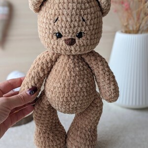 K&ouml;nnte beinhalten: Ein brauner geh&auml;kelter Teddyb&auml;r mit schwarzen Augen und einer braunen Nase. Der B&auml;r steht auf einer wei&szlig;en Oberfl&auml;che.