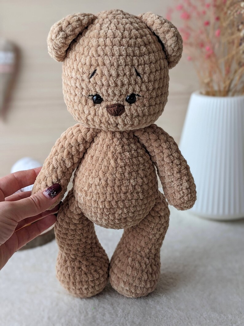 K&ouml;nnte beinhalten: Ein brauner geh&auml;kelter Teddyb&auml;r mit schwarzen Augen und einer braunen Nase. Der B&auml;r steht auf einer wei&szlig;en Oberfl&auml;che.