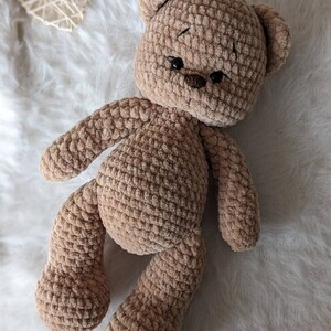K&ouml;nnte beinhalten: Ein brauner geh&auml;kelter Teddyb&auml;r mit schwarzen Augen und einer braunen Nase. Der B&auml;r liegt auf einer wei&szlig;en, flauschigen Oberfl&auml;che.