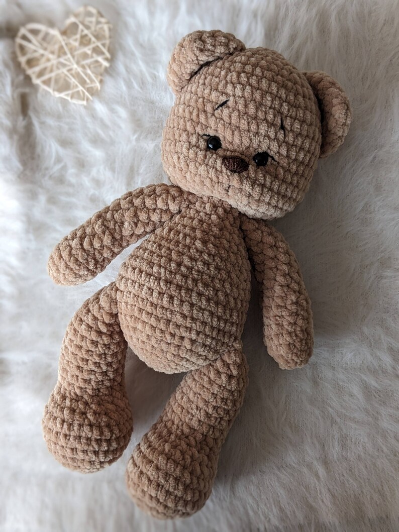 K&ouml;nnte beinhalten: Ein brauner geh&auml;kelter Teddyb&auml;r mit schwarzen Augen und einer braunen Nase. Der B&auml;r liegt auf einer wei&szlig;en, flauschigen Oberfl&auml;che.