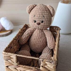 K&ouml;nnte beinhalten: Ein brauner geh&auml;kelter Teddyb&auml;r sitzt in einem geflochtenen Weidenkorb. Der B&auml;r hat gestickte Augen und eine gen&auml;hte Nase.