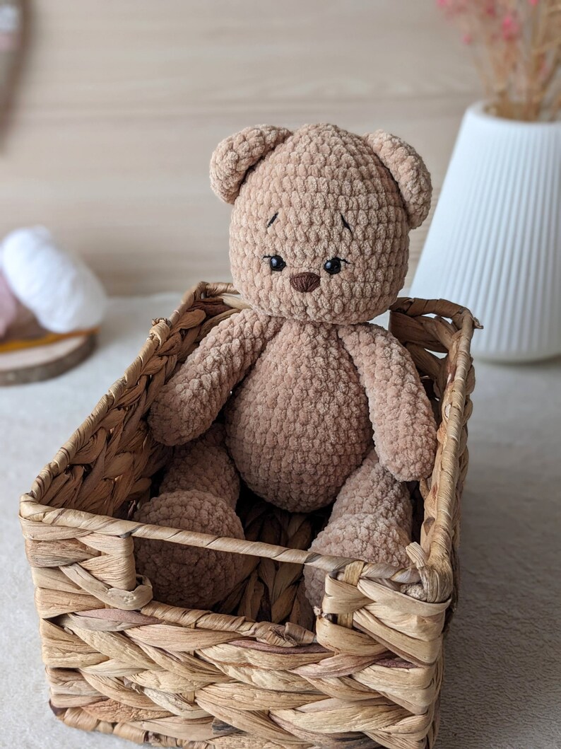 K&ouml;nnte beinhalten: Ein brauner geh&auml;kelter Teddyb&auml;r sitzt in einem geflochtenen Weidenkorb. Der B&auml;r hat gestickte Augen und eine gen&auml;hte Nase.