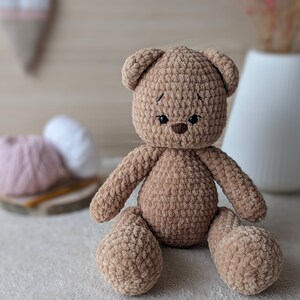 K&ouml;nnte beinhalten: Ein brauner geh&auml;kelter Teddyb&auml;r mit schwarzen Augen und einer braunen Nase. Der B&auml;r sitzt auf einer wei&szlig;en Oberfl&auml;che.