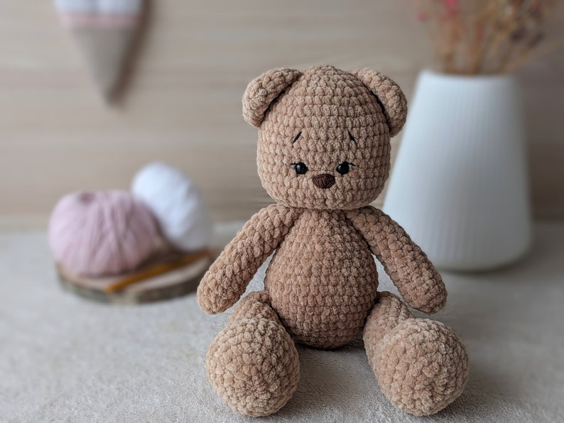 K&ouml;nnte beinhalten: Ein brauner geh&auml;kelter Teddyb&auml;r mit schwarzen Augen und einer braunen Nase. Der B&auml;r sitzt auf einer wei&szlig;en Oberfl&auml;che.