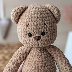 K&ouml;nnte beinhalten: Ein brauner geh&auml;kelter Teddyb&auml;r mit schwarzen Augen und einer braunen Nase. Der B&auml;r ist aus weichem, pl&uuml;schigen Garn gefertigt.
