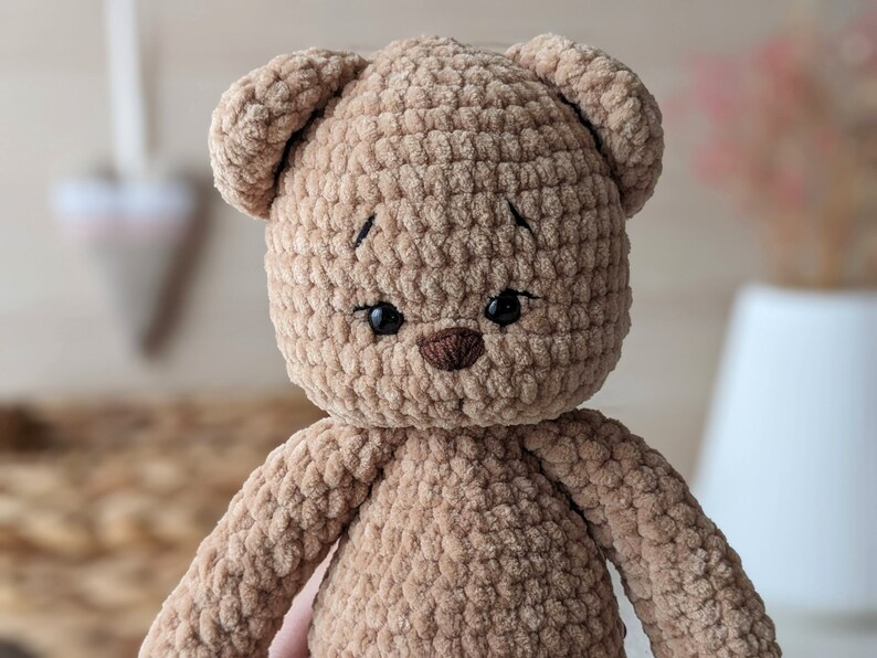 K&ouml;nnte beinhalten: Ein brauner geh&auml;kelter Teddyb&auml;r mit schwarzen Augen und einer braunen Nase. Der B&auml;r ist aus weichem, pl&uuml;schigen Garn gefertigt.