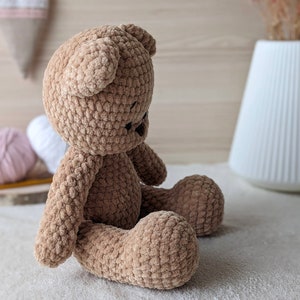 K&ouml;nnte beinhalten: Ein brauner geh&auml;kelter Teddyb&auml;r, der auf einer wei&szlig;en Oberfl&auml;che sitzt. Der B&auml;r hat eine weiche, flauschige Textur und ist aus braunem Garn gefertigt.