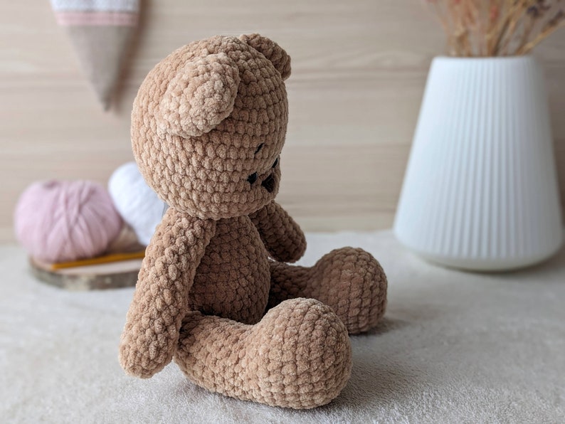 K&ouml;nnte beinhalten: Ein brauner geh&auml;kelter Teddyb&auml;r, der auf einer wei&szlig;en Oberfl&auml;che sitzt. Der B&auml;r hat eine weiche, flauschige Textur und ist aus braunem Garn gefertigt.