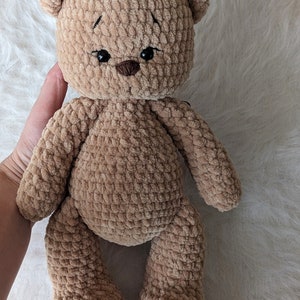 K&ouml;nnte beinhalten: Ein brauner geh&auml;kelter Teddyb&auml;r mit schwarzen Augen und einer braunen Nase. Der B&auml;r sitzt auf einer wei&szlig;en, flauschigen Oberfl&auml;che.