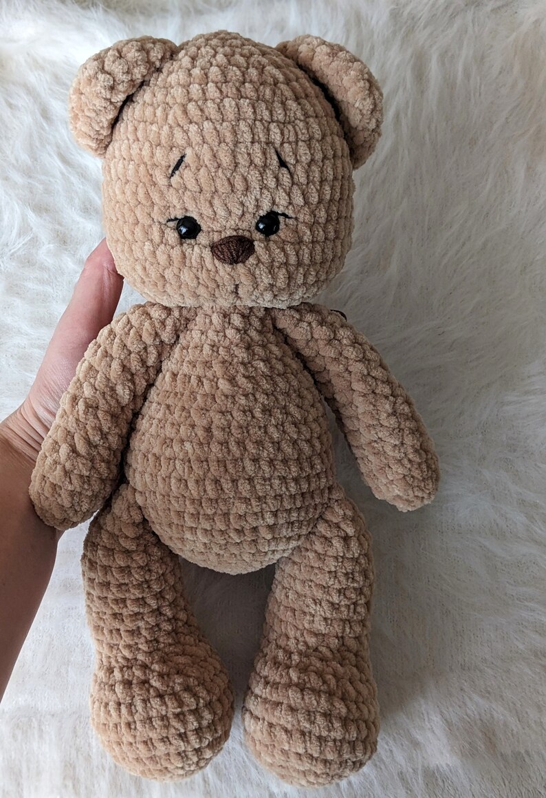 K&ouml;nnte beinhalten: Ein brauner geh&auml;kelter Teddyb&auml;r mit schwarzen Augen und einer braunen Nase. Der B&auml;r sitzt auf einer wei&szlig;en, flauschigen Oberfl&auml;che.