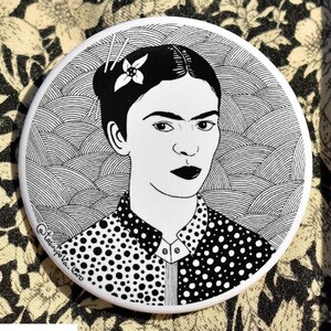 May include: A black and white illustration of Frida Kahlo with a flower in her hair. The background is a pattern of overlapping circles and lines. The image is printed on a round white button.