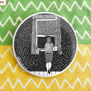 Könnte beinhalten: Ein runder, schwarz-weißer Button mit einer detaillierten Illustration einer Person, die aus einem Fenster auf eine verschneite Szene blickt. Der Button hat einen gepunkteten Rand und den Text "Tasirupeka Haro".