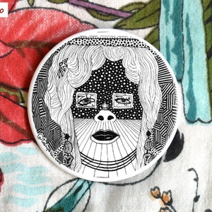 May include: Black and white line art illustration of a woman's face with geometric patterns. The woman has long hair and is wearing a necklace. The background is a white circle.