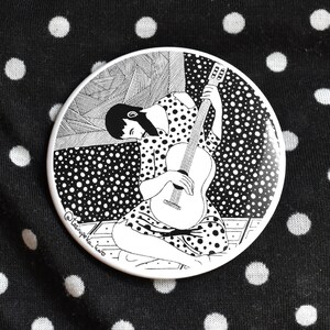 May include: A circular button with a black and white illustration of a person playing a guitar. The person is seated and wearing a polka-dot dress. The button is set against a black fabric with white polka dots.