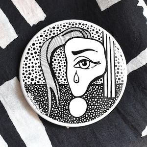 Op de afbeelding: Een ronde zwarte en witte knop met een gestileerde illustratie van een huilend vrouwen gezicht. De achtergrond is een zwart-wit stippenpatroon.