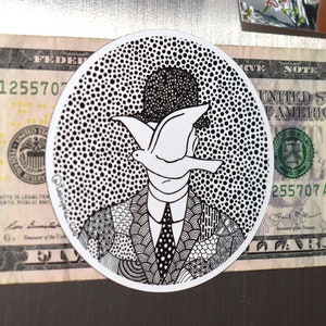 Peut inclure: Un autocollant rond avec une illustration en noir et blanc d'une personne avec une colombe à la place du visage, placé sur un billet de cinq dollars. L'autocollant est sur une surface métallique.