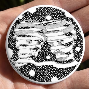 May include: A black and white button featuring a design of two faces wrapped in ribbons. The background is a black and white polka dot pattern.