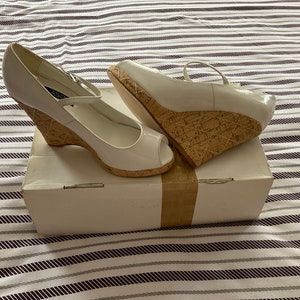 Puede incluir: Un par de sandalias de cuña de charol blanco con diseño de punta abierta. Los tacones de cuña son de corcho y las sandalias tienen una correa en el tobillo. Las sandalias están sobre una caja blanca.