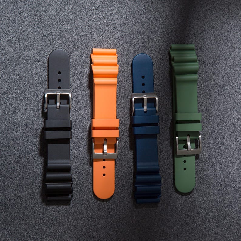 May include: Four different coloured watch straps, black, orange, blue, and green, are displayed against a grey background. The straps are made of a rubber-like material and have a buckle fastening.