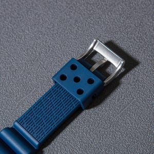May include: Close-up of a blue watch strap with a silver buckle. The strap has a textured surface and a row of holes.