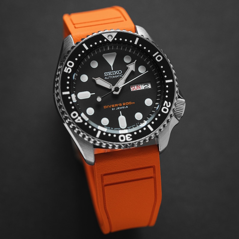 Op de afbeelding: Een zwart en zilveren Seiko automatisch horloge met een oranje rubberen band. De wijzerplaat van het horloge heeft een zwarte achtergrond met witte markeringen en een datumscherm dat "SUN 2" aangeeft. De bezel is zwart met witte markeringen en geeft "DIVER'S 200m 21 JEWELS" aan.