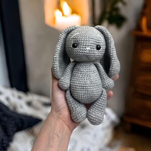 Puede incluir: Un juguete de conejo de crochet gris con ojos negros y detalles blancos en las orejas. El conejo está siendo sostenido por una mano.