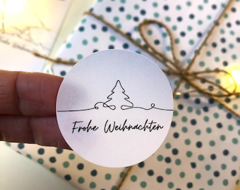 May include: A wrapped gift with white and blue polka dot paper, tied with twine. A round sticker with a minimalist Christmas tree design and the words "Frohe Weihnachten" is held in front of the gift.