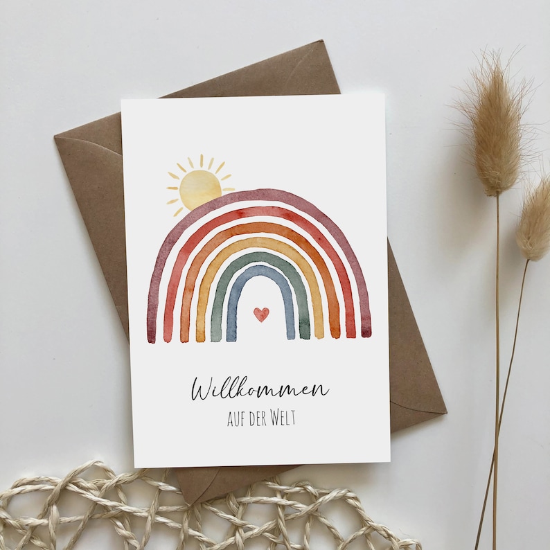 Postkarte auf braunem Umschlag aus Kraftpapier mit Motiv: Regenbogen mit aufgehender Sonne. Darunter der Text: Willkommen auf der Welt.