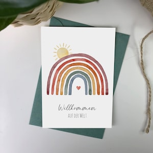 Postkarte auf grünem Umschlag aus Kraftpapier mit Motiv: Regenbogen mit aufgehender Sonne. Darunter der Text: Willkommen auf der Welt.