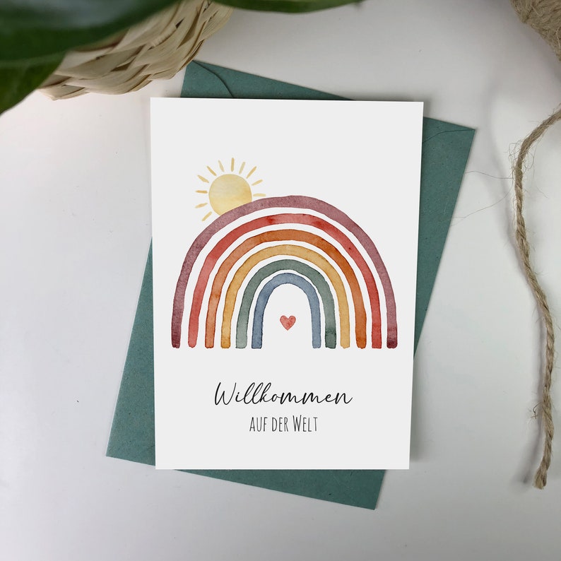Postkarte auf grünem Umschlag aus Kraftpapier mit Motiv: Regenbogen mit aufgehender Sonne. Darunter der Text: Willkommen auf der Welt.
