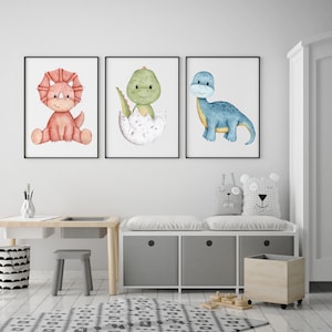 May include: Three watercolor dinosaur prints in white frames. The prints feature a red dinosaur, a green dinosaur in an egg, and a blue dinosaur. The prints are hanging on a white wall above a white bench with two gray cushions and a wooden storage bin.