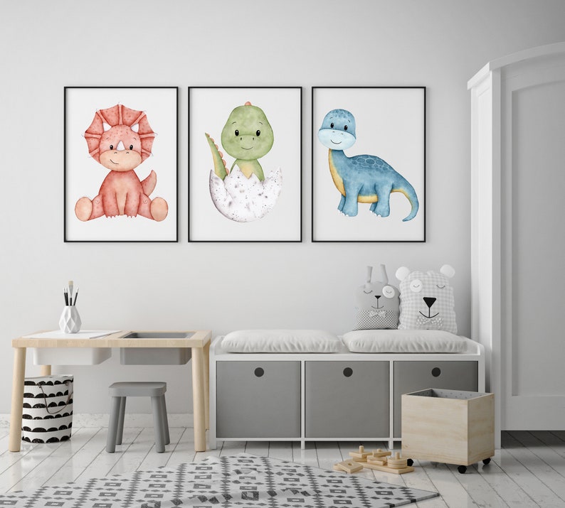 May include: Three watercolor dinosaur prints in white frames. The prints feature a red dinosaur, a green dinosaur in an egg, and a blue dinosaur. The prints are hanging on a white wall above a white bench with two gray cushions and a wooden storage bin.