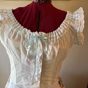 May include: A white cotton blouse with a lace trim and a blue ribbon bow. The blouse has a fitted bodice and short sleeves.