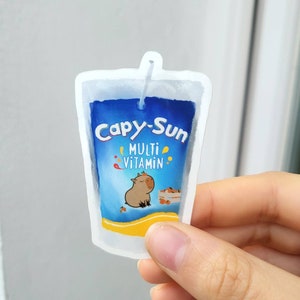 Op de afbeelding: Een gestanste sticker in de vorm van een sapzakje met een blauwe achtergrond, voorzien van de tekst Capy-Sun en MULTI VITAMIN. Het ontwerp bevat een kleine bruine capibara-illustratie, een kistje met sinaasappels en een wit rietje bovenaan.