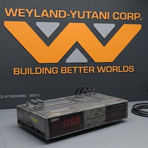 Op de afbeelding: Een rechthoekig, verweerd grijs elektronisch apparaat met een digitaal display dat 1:00 aangeeft, voorzien van een zwarte stroomkabel en een logo aan de muur met de tekst WEYLAND-YUTANI CORP. BUILDING BETTER WORLDS en OFFICE OF PERSONNEL - DECK C.