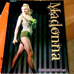 May include: A book cover with a photo of Madonna in a green and white striped costume. The title "Madonna" is in gold lettering on a black and gold striped background. The book is by Daryl Easlea & Eddi Fiegel.
