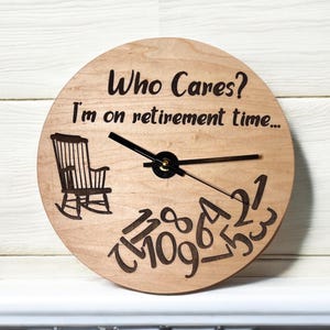 May include: A round wooden clock with the text "Who Cares? I'm on retirement time..." and a rocking chair illustration. The clock hands are black, and the numbers are arranged in a unique, stylized font.