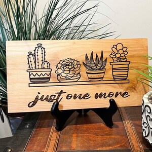 May include: Wooden sign with a burnt-in design of four potted succulents on a shelf with the text "just one more".