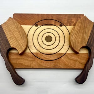 May include: A rectangular wooden serving board featuring a central light-colored wood inlay with concentric dark circles and two small dots. Two axe-shaped wooden handles with dark and light wood grain patterns are attached to the sides of the board.