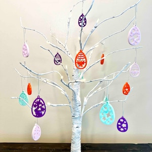 May include: A white birch tree with a white base, decorated with colorful Easter egg ornaments. The ornaments are made of plastic and have a variety of designs, including flowers, butterflies, and geometric patterns. The tree is lit with white lights.