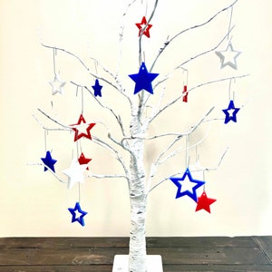 May include: A white birch tree with a white base and red, white, and blue star ornaments hanging from the branches.