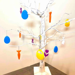 May include: A white decorative tree with a square base, featuring hanging ornaments in various shapes including blue eggs, orange carrots, yellow eggs, purple flowers, and white rabbits, all suspended by thin silver wires from the bare branches.