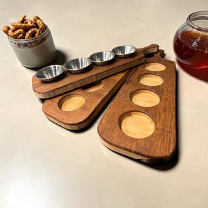 May include: Two rectangular wooden serving boards with rounded edges, each featuring four circular cutouts. One board holds four small, round, silver-colored metal bowls. A glass jar filled with pretzels and a glass jar of honey are nearby.