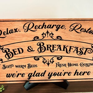 May include: A wooden sign with the text "Relax, Recharge, Restore Bed & Breakfast Soft Warm Beds Fresh Home Cooking we're glad you're here". The sign is painted in black and has a decorative scroll design.
