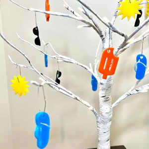 May include: A white birch tree with a white base is decorated with various summer-themed ornaments. The ornaments include a yellow sun, blue flip-flops, orange popsicles, and black sunglasses.